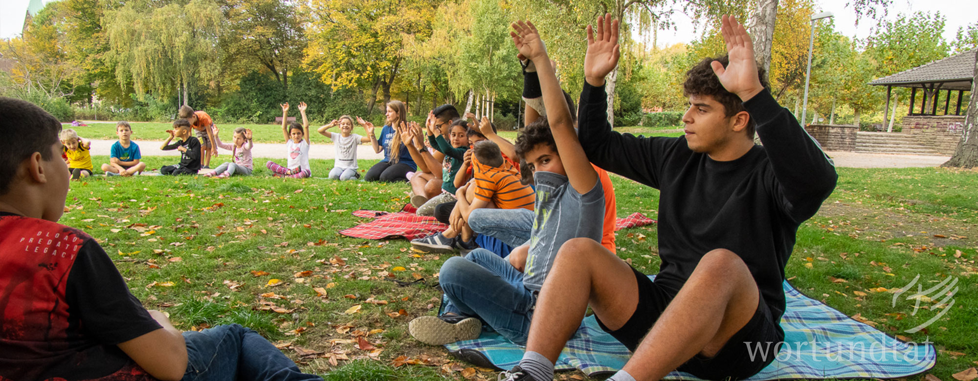 junger Mann sitzt mit Kindern auf Wiese - alle haben Hände erhoben - ehrenamtlich helfen in Dortmund
