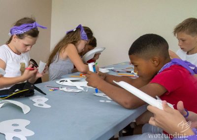 Mehrere Kinder sitzen an einem Tisch und basteln mit Schere Kleber, Papier, und Stiften kleine Gesichtsmasken, die sie verzieren