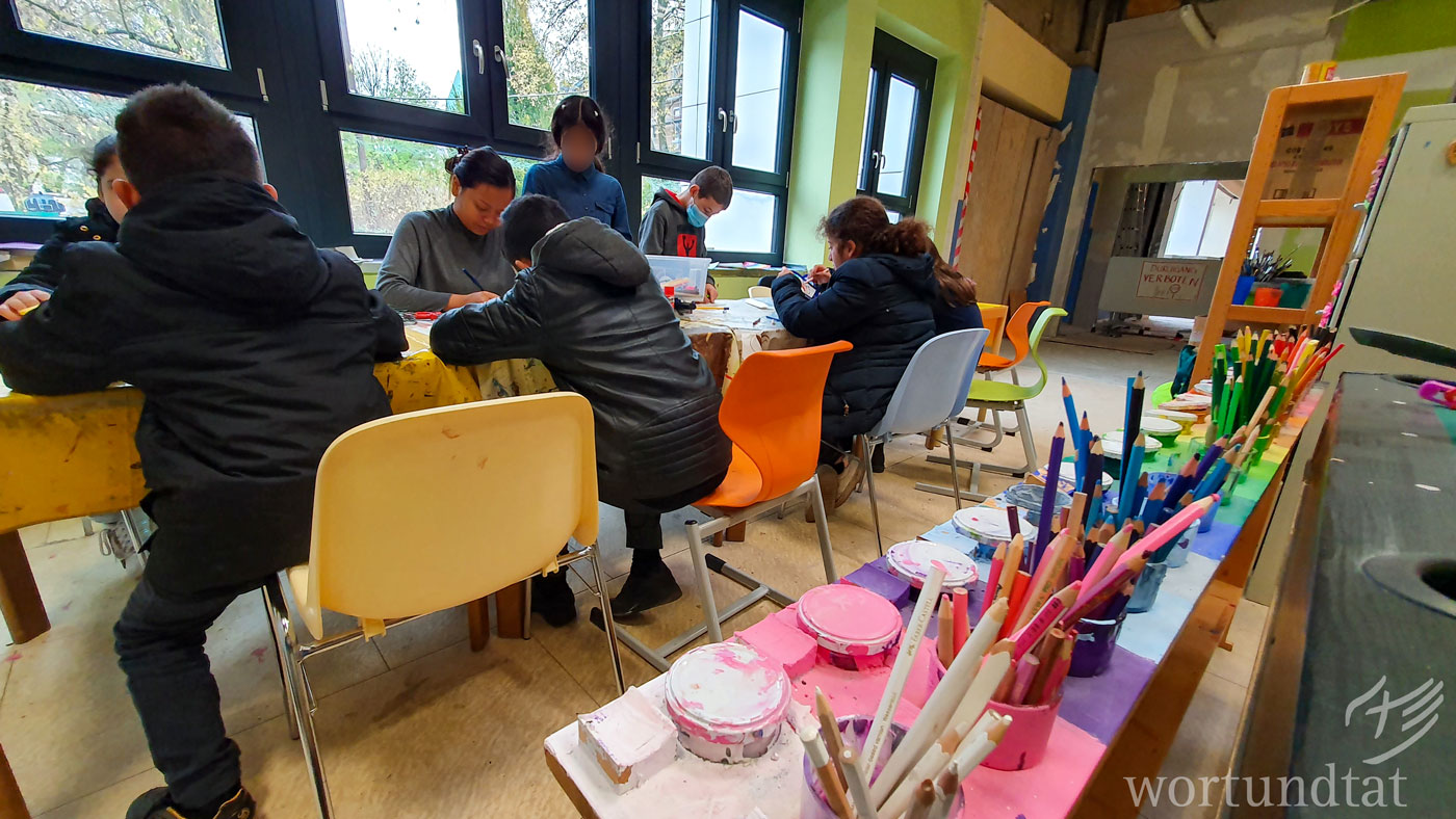 Kinder sitzen an einem Tisch und basteln - im Vordergrund farbige Buntstifte