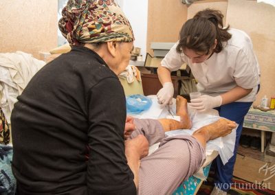 Nurse cares for an open leg of a patient sitting in simply furnished room